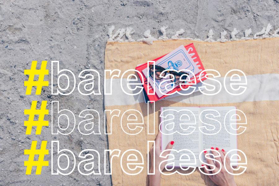 på en strand på et tæppe en stak bøger med solbriller ovenpå et par hænder der holder en bog